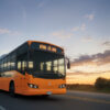 夕焼けの中を走るオレンジ色のバスが、まっすぐな道路上に映える光景。 / An orange bus driving along a straight road, glowing in the warm light of sunset.