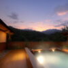 夕暮れの山々を望む木造の露天風呂と静かな温泉宿の一角。 / An open-air hot spring bath with wooden decking, overlooking quiet mountains at dusk.