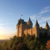 青空の下、山の上にそびえる壮麗な塔付きの城の風景。 / A majestic castle with towers standing atop a hill under a clear blue sky.