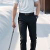 雪景色の中、白いポロシャツと黒いパンツ姿で手に棒を持ち、雪上を歩く男性。/A man in a white polo shirt and black pants walking on snow, holding a stick in a snowy landscape.