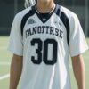 白のラクロスユニフォームを着た青年がフィールドでスティックを肩に微笑む A young man in a white lacrosse uniform smiling with his stick resting on his shoulder on the field
