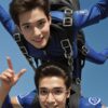 青空を背景に2人の青年がスカイダイビング中に笑顔で手を振る躍動感あふれる瞬間 Two young men smiling and waving mid-skydive against a bright blue sky