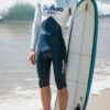 白と黒のウェットスーツを着た青年が砂浜でサーフボードの横に立つ A young man in a white and black wetsuit standing on the beach beside a surfboard