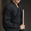 黒い剣道着を着た若い男性が竹刀を持ち、道場内でこちらを見つめている。 / A young man in a black kendo uniform is holding a bamboo sword and looking at the camera inside a dojo.