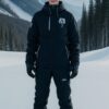 黒のスノーボードウェアを着た若い男性が雪山でボードに乗って正面を見ている。 / A young man in black snowboard wear is standing on a snowboard in the snow, looking forward.