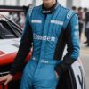 青いレーシングスーツを着た若い男性がレーシングカーの横で微笑んで立っている。 / A young man in a blue racing suit is smiling while standing next to a race car.