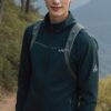 濃緑のハイキングウェアを着た若い男性が山道でリュックを背負い、微笑んでいる。 / A young man in dark green hiking wear is smiling while carrying a backpack on a mountain trail.