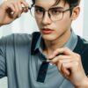 Young man in a gray polo shirt sitting by the window, adjusting his glasses with one finger （灰色のポロシャツを着た若い男性が窓際に座り、片手の指でメガネを調整している）