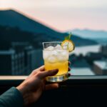 氷の入ったグラスを片手で持ち、夕暮れの街並みを背景にした構図 / A hand holding an iced drink with a cityscape at sunset in the background