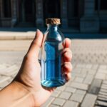 片手で青い液体の入ったボトルを持ち、石畳の道を背景にした構図 / A hand holding a bottle with blue liquid, with cobblestone pavement in the background
