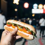 片手で持ったホットドッグを夜の通りで掲げる構図 / A hotdog held up with one hand at night on a busy street