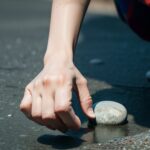 濡れた地面で片手を伸ばして石を拾い上げようとしている様子 / A person reaching down with one hand to pick up a pebble on wet ground