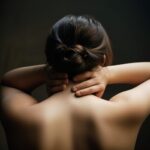 まとめ髪の女性が両手を首に当てて肩の緊張をほぐしている様子の写真。 / A photo of a woman with tied-up hair relaxing her shoulders by placing both hands on her neck.