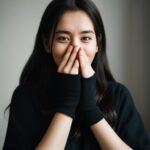 黒い服を着た女性が両手で口を押さえて微笑んでいる写真。 / A photo of a woman in black clothing gently smiling while covering her mouth with both hands.