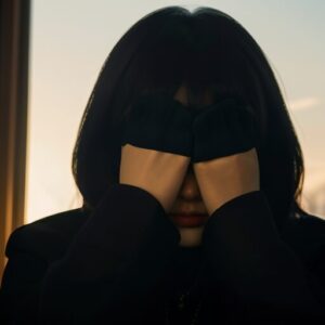 暗がりの中でサングラスをかけ、両手でさらに目を覆う女性の写真。 / A photo of a woman wearing sunglasses and covering her eyes with both hands in dim lighting.