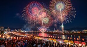 川沿いの都市の夜景を背景に大輪の打ち上げ花火が夜空に広がり、水面に光が映り込む中、提灯の灯る屋台と大勢の観客が見上げる夏祭りの光景（Gemini（Nano Banana）で生成）
