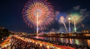 夜空に大輪の打ち上げ花火が広がり、川面に光が映る中、提灯と屋台が並ぶ川沿いを大勢の観客が埋める夏祭りの夜景（Gemini（Nano Banana）で生成）