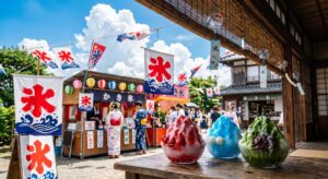 「氷」の旗や提灯が飾られた明るい夏の街並みで、いちご・ブルーハワイ・宇治金時のかき氷が屋台の店先に並び、浴衣姿の人々が行き交う風景(Gemini(Nano Banana)で生成)